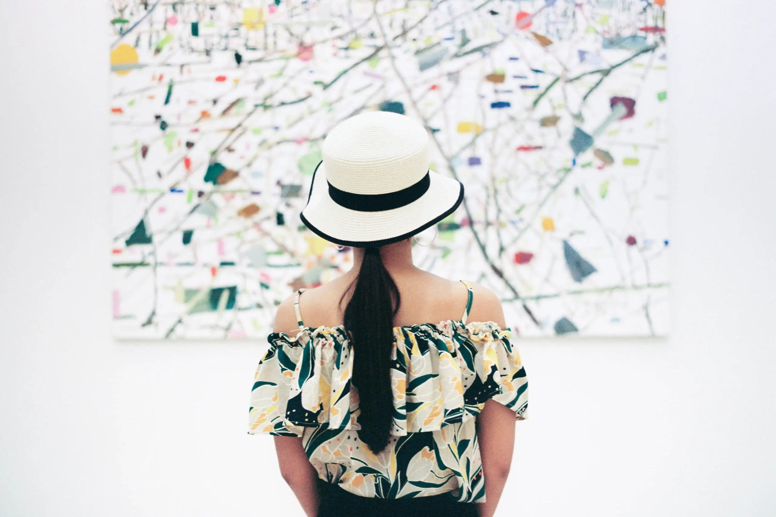 Woman observing abstract art in a gallery, wearing a floral dress and a white hat